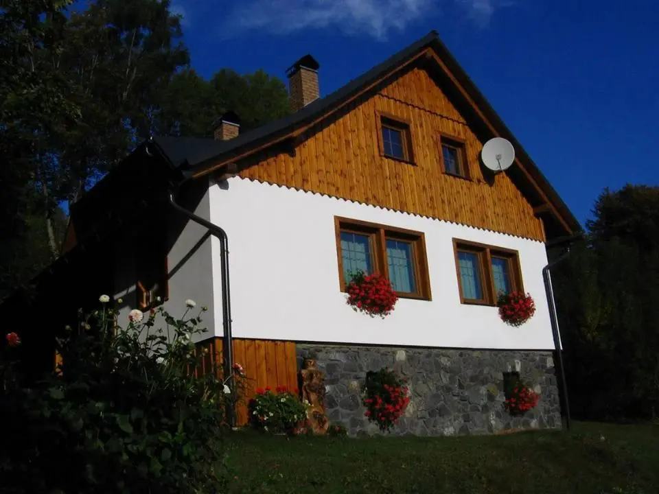 Detail dokončené roubenky Deštné s bílou fasádou, dřevěným štítem a pelargoniemi v oknech