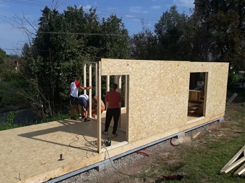 Montáž rámové stěny dřevostavby s otvorem pro vchodové dveře na základové desce